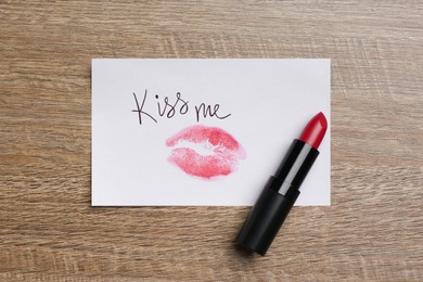 Photo of Paper card with lip print, phrase Kiss Me and lipstick on wooden table, top view