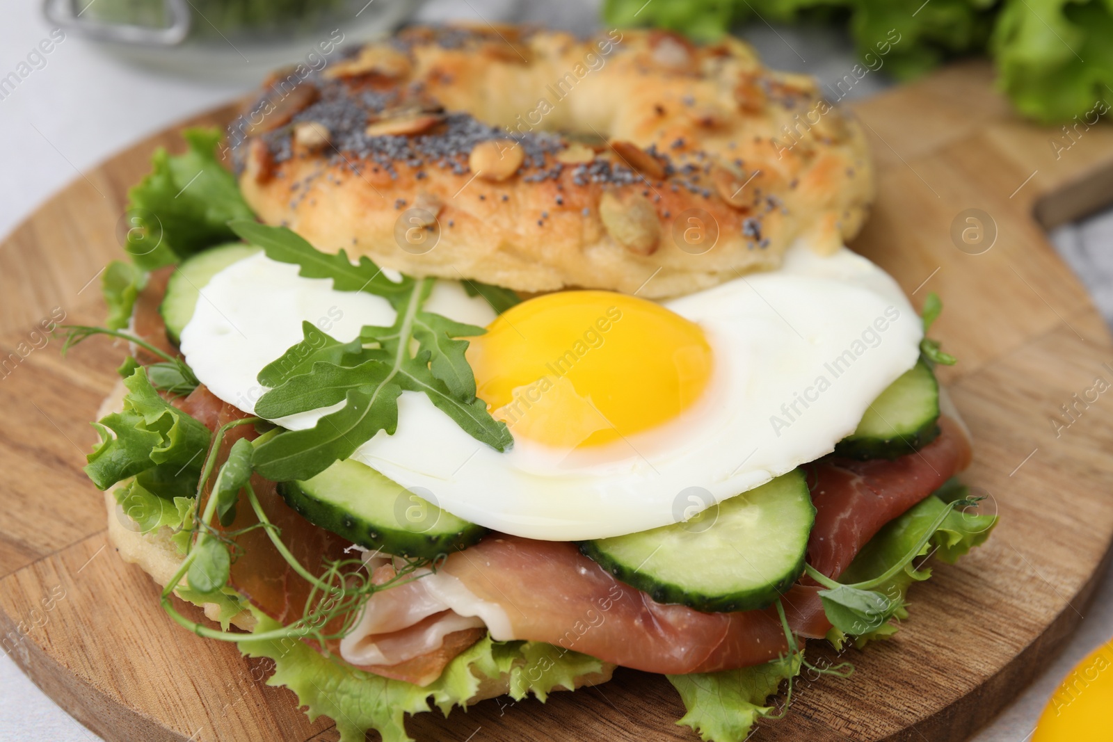 Photo of Tasty bagel with cured ham, egg, cucumber and salad mix on table, closeup