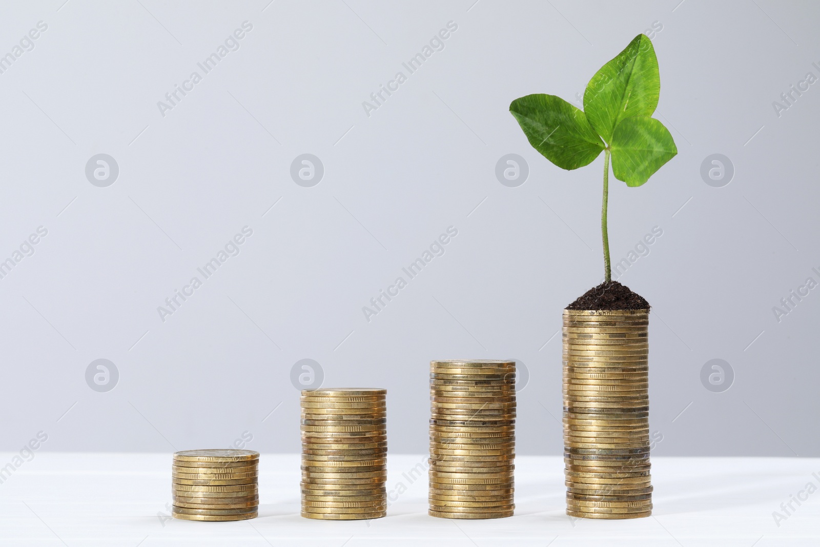 Photo of Stacks of coins with green sprout on white table against light grey background, space for text. Investment concept