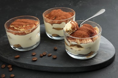 Photo of Delicious tiramisu in glasses, spoon and coffee beans on black table