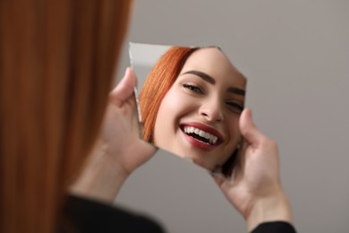 Smiling young woman looking at herself in broken mirror on light grey background, closeup