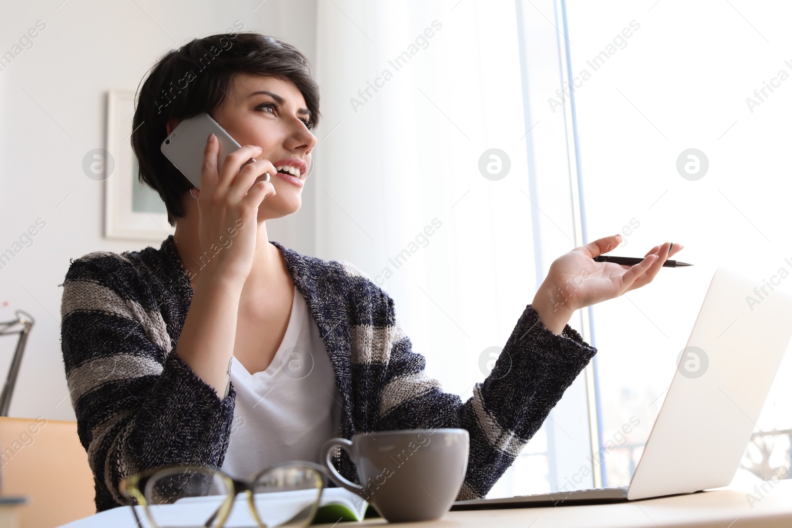 Photo of Young woman talking on mobile phone while working with laptop at desk. Home office