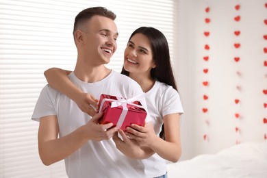 Photo of Girlfriend presenting gift to her boyfriend indoors, space for text. Valentine`s day celebration
