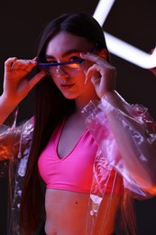 Photo of Fashionable portrait of beautiful woman wearing transparent coat and glasses on dark background in neon lights