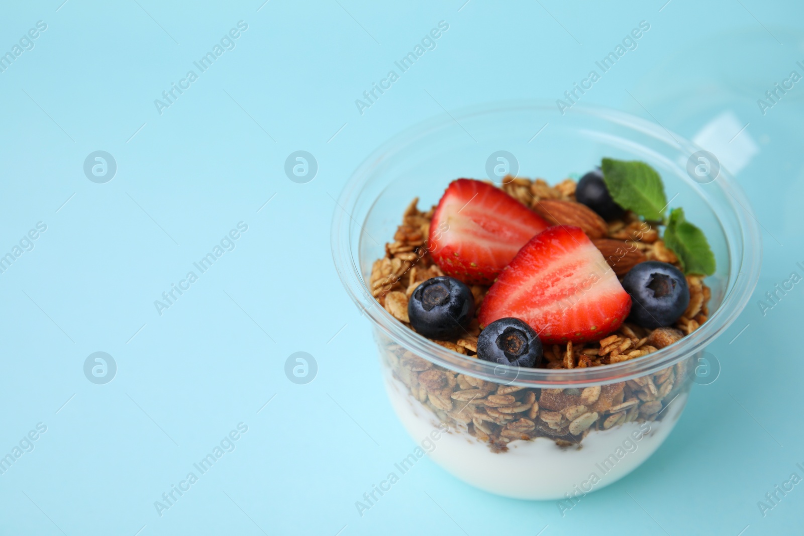 Photo of Tasty granola with berries, nuts and yogurt in plastic cup on light blue background, closeup. Space for text