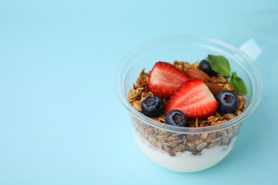 Photo of Tasty granola with berries, nuts and yogurt in plastic cup on light blue background, closeup. Space for text