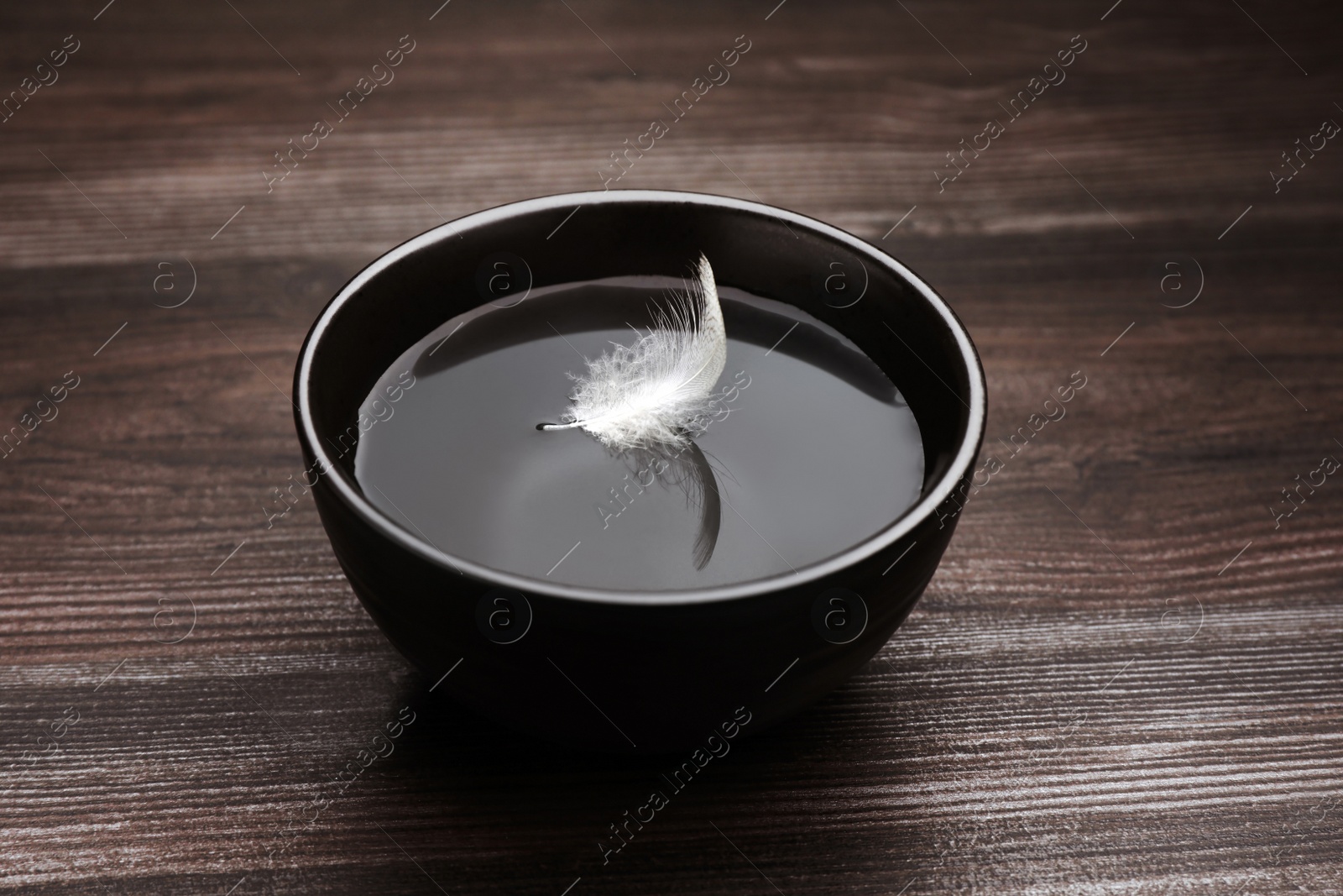 Photo of Black bowl with water and white feather on dark wooden table