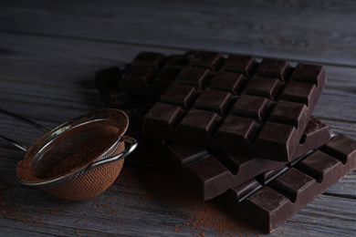 Photo of Tasty dark chocolate bars and sieve with cocoa powder on wooden table