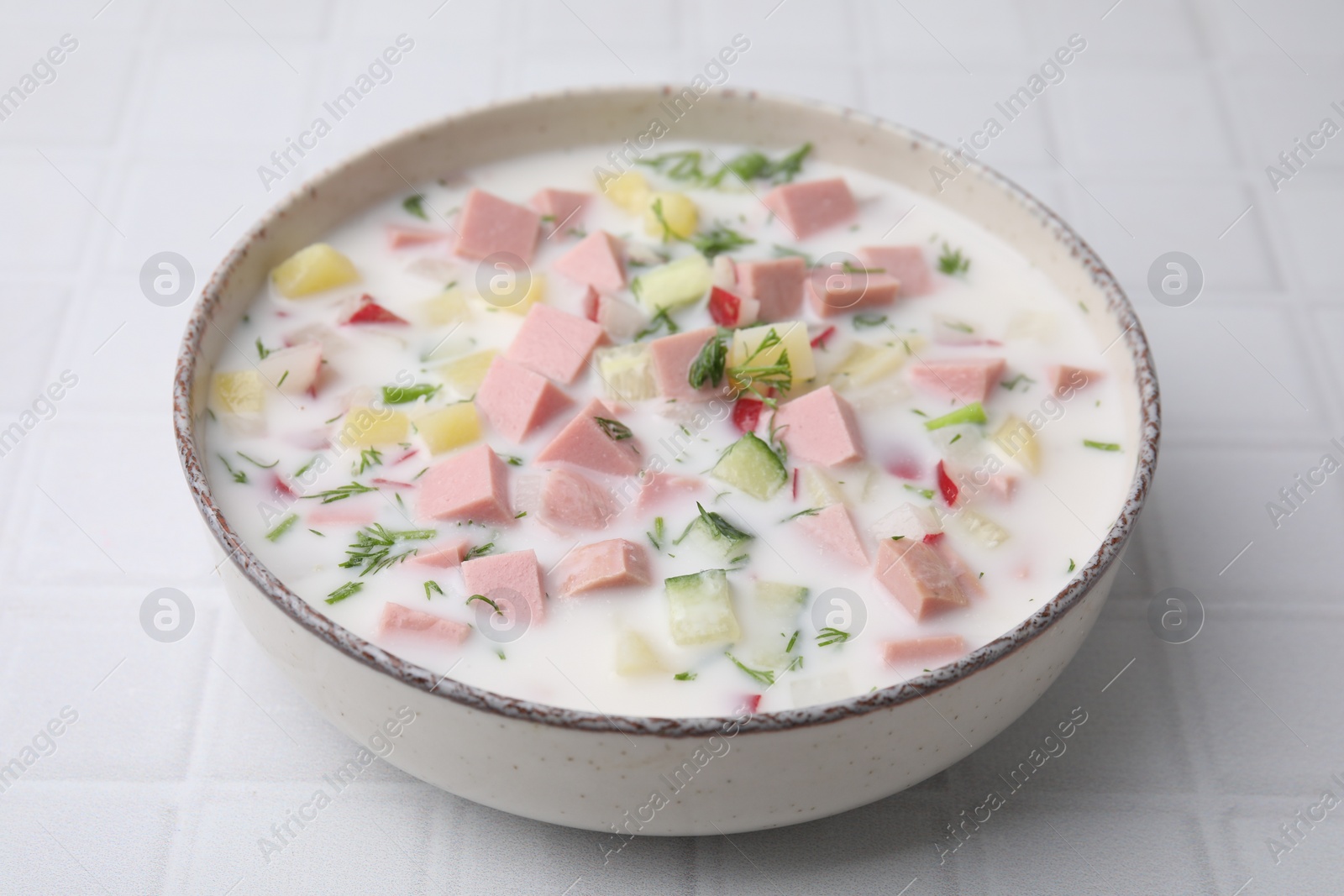 Photo of Delicious cold summer soup (okroshka) with boiled sausage in bowl on white tiled table, closeup
