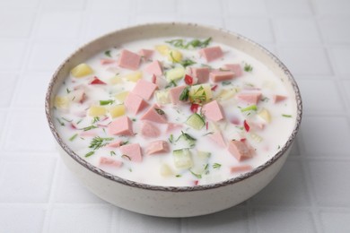 Delicious cold summer soup (okroshka) with boiled sausage in bowl on white tiled table, closeup