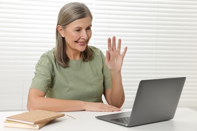 Photo of Happy woman waving hello during video call at table indoors