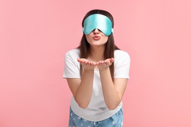 Woman in pyjama and sleep mask blowing kiss on pink background