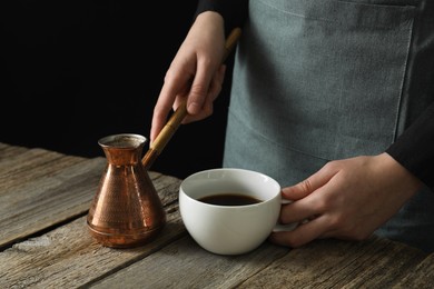 Turkish coffee. Woman with cezve and cup of freshly brewed beverage at wooden table against black background, closeup