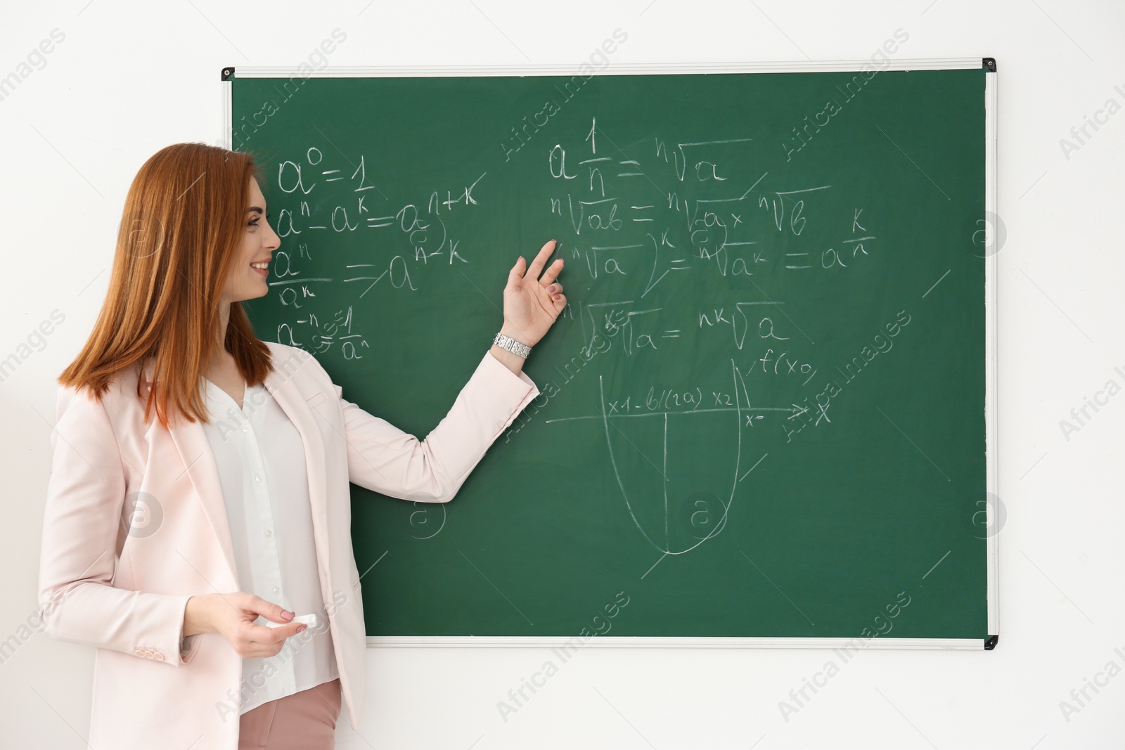 Photo of Beautiful young teacher explaining math formulas written on blackboard in classroom