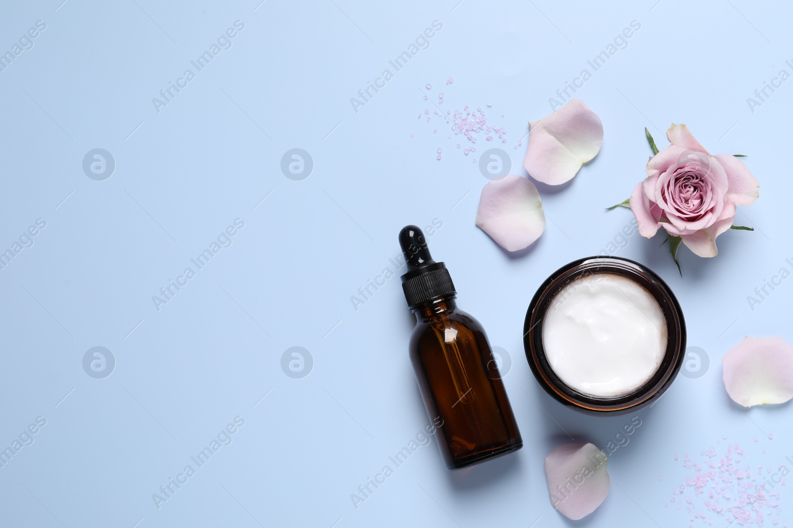 Photo of Bottle of cosmetic serum, cream, flower and petals on light blue background, flat lay. Space for text