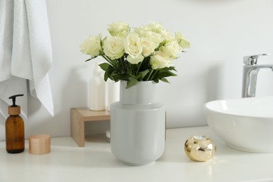 Vase with beautiful white roses and toiletries near sink in bathroom