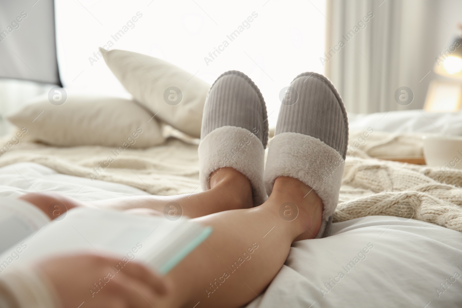 Photo of Woman in soft comfortable slippers reading book on bed at home, closeup