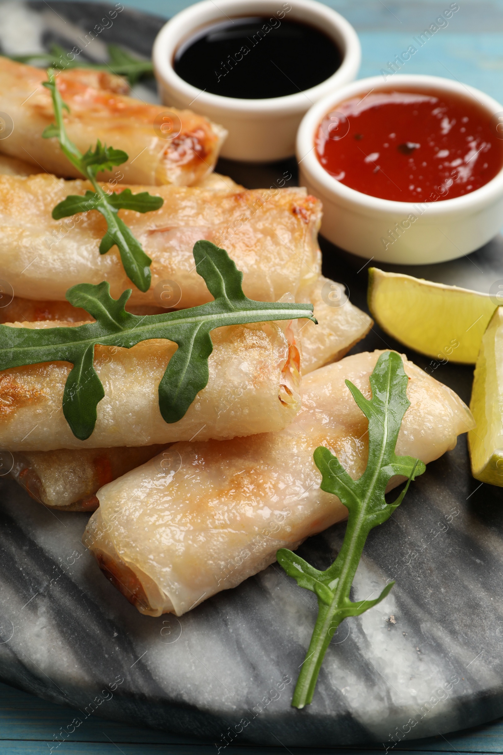 Photo of Tasty fried spring rolls, arugula, lime and sauces on light blue table, closeup
