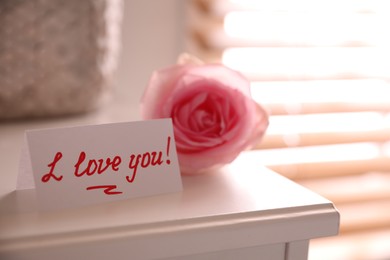 Photo of Note with handwritten text I Love You near pink rose on white table in room, closeup