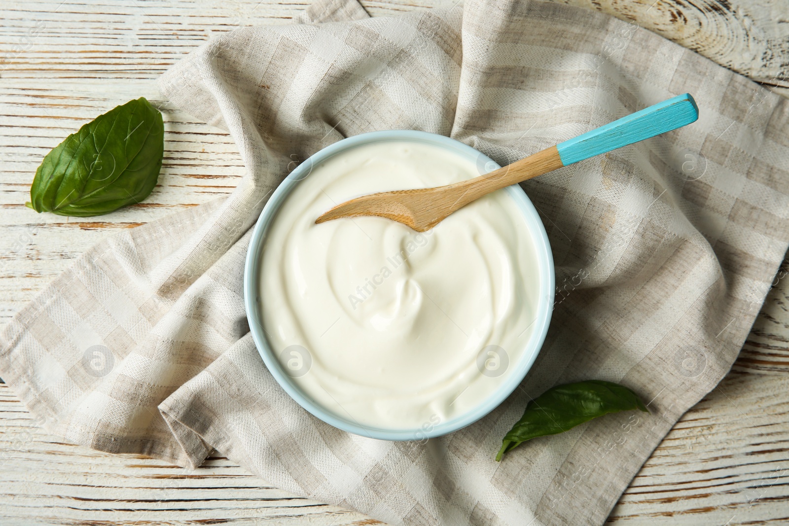 Photo of Bowl of sour cream with spoon and napkin on white wooden table, flat lay