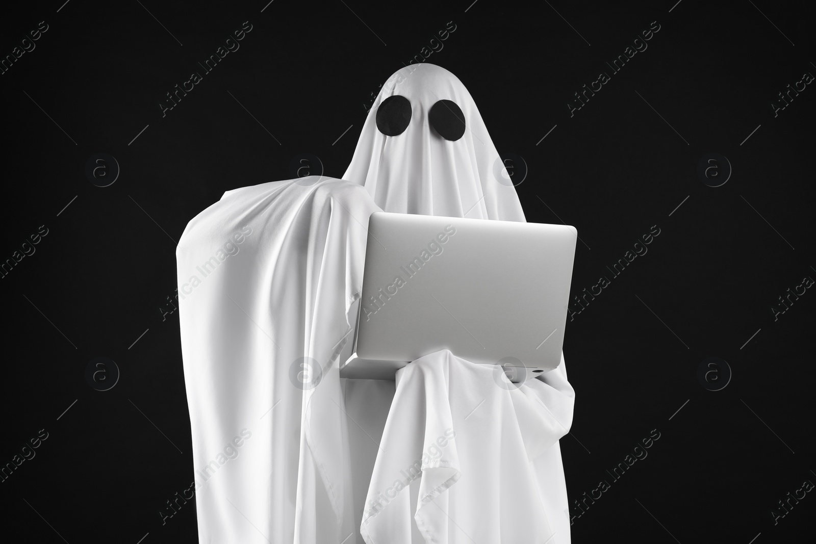 Photo of Creepy ghost. Person in white sheet with laptop on black background