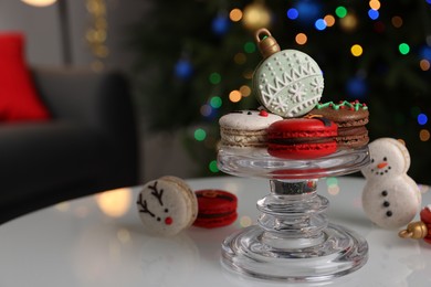 Beautifully decorated Christmas macarons on white table against blurred festive lights, space for text