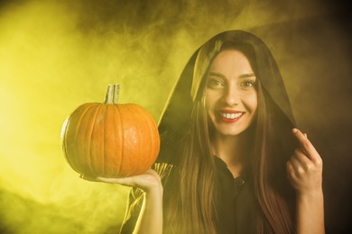 Photo of Young woman wearing witch costume with pumpkin in smoke cloud on dark background. Halloween party