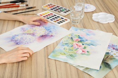 Woman holding painting of flowers near watercolor at wooden table, closeup. Creative artwork