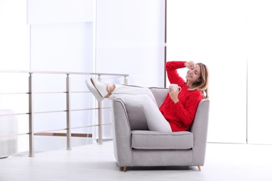 Attractive young woman with cup of tea in armchair indoors. Space for text