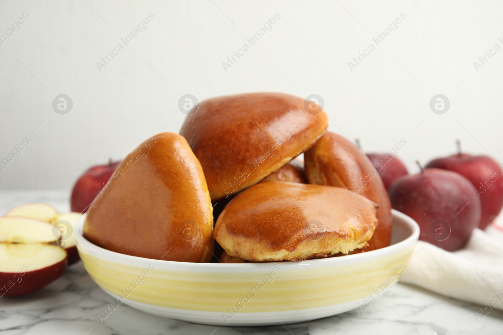 Photo of Delicious baked apple pirozhki in bowl and fruits on white marble table