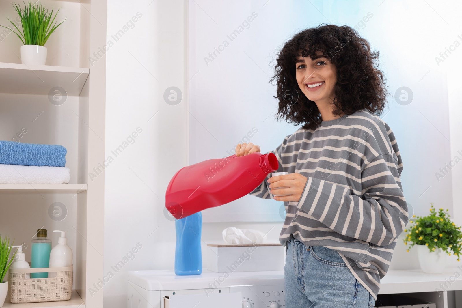 Photo of Happy woman pouring laundry detergent into cap near washing machine indoors