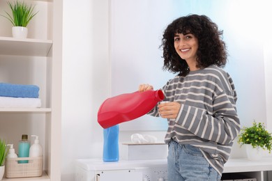 Happy woman pouring laundry detergent into cap near washing machine indoors