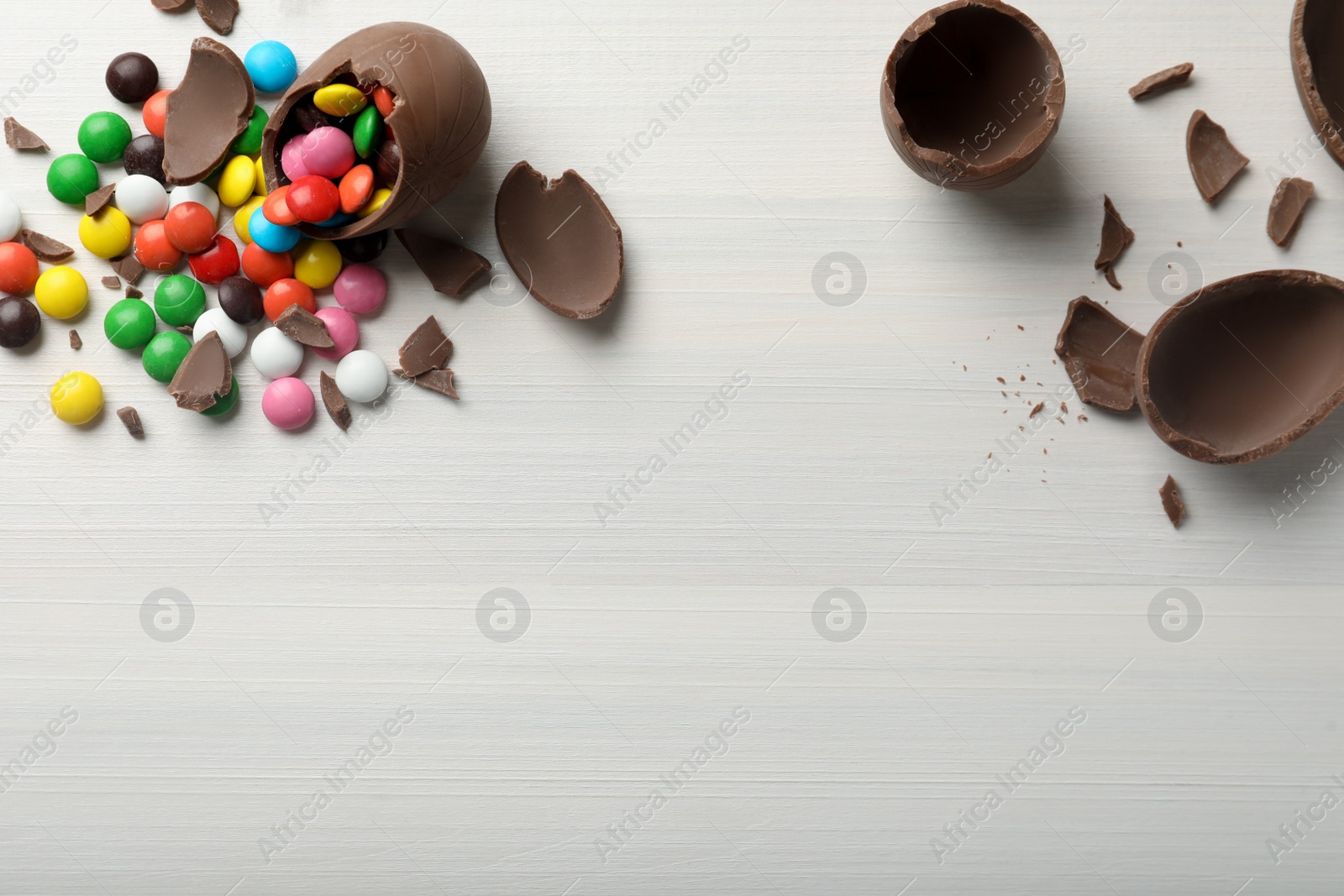 Photo of Broken chocolate eggs and colorful candies on white wooden table, flat lay. Space for text