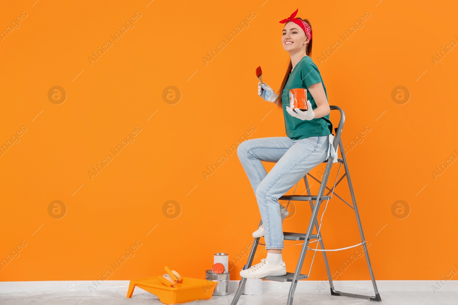 Photo of Happy designer with painting equipment sitting on folding ladder near freshly painted orange wall indoors, space for text