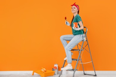 Photo of Happy designer with painting equipment sitting on folding ladder near freshly painted orange wall indoors, space for text