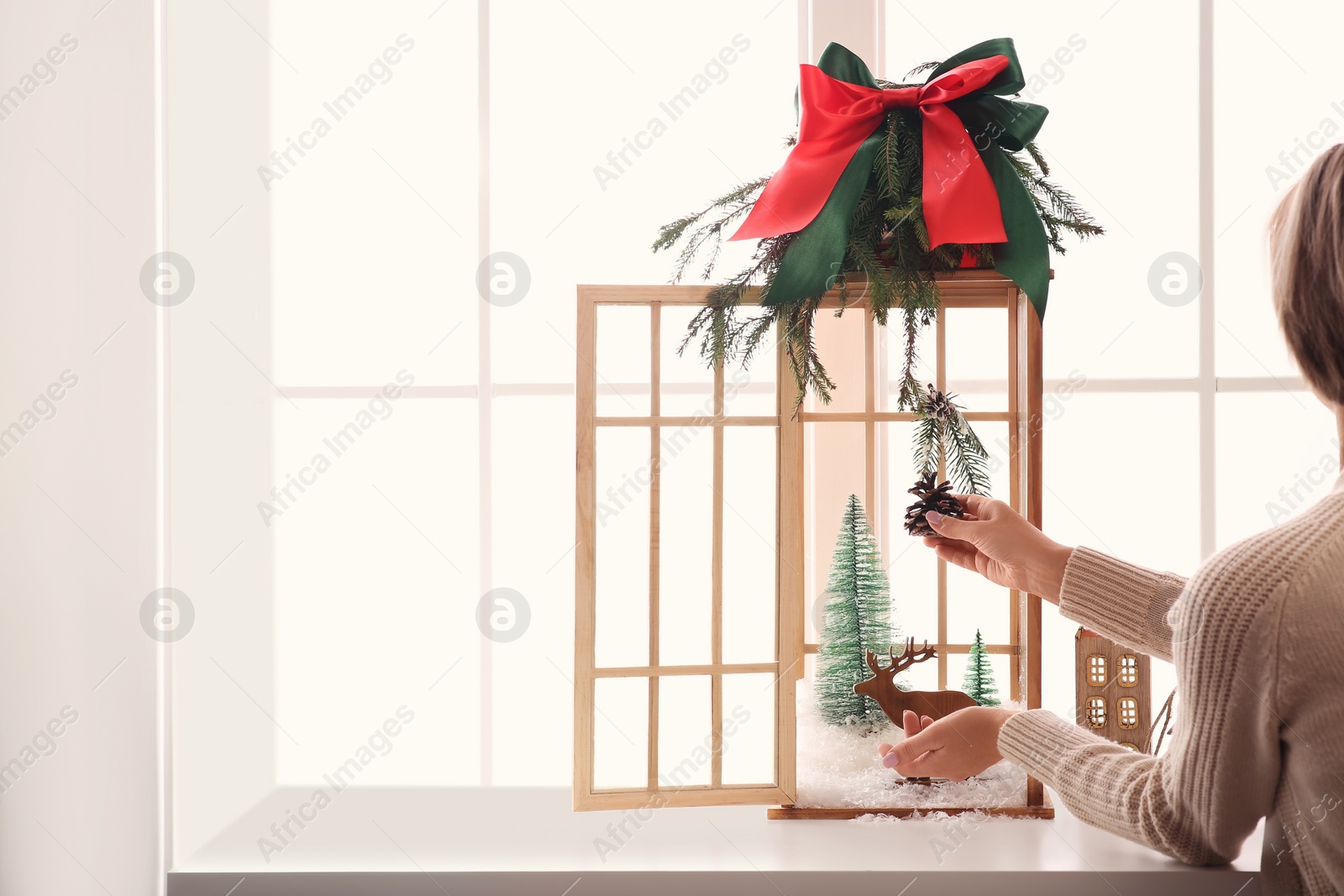 Photo of Woman creating Christmas composition inside of vintage wooden lantern on window sill indoors, closeup. Space for text