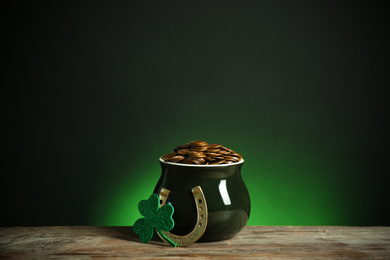 Photo of Pot with gold coins, horseshoe and clover on wooden table against dark background. St. Patrick's Day celebration