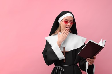 Photo of Surprised woman in nun habit and sunglasses reading Bible against pink background. Space for text