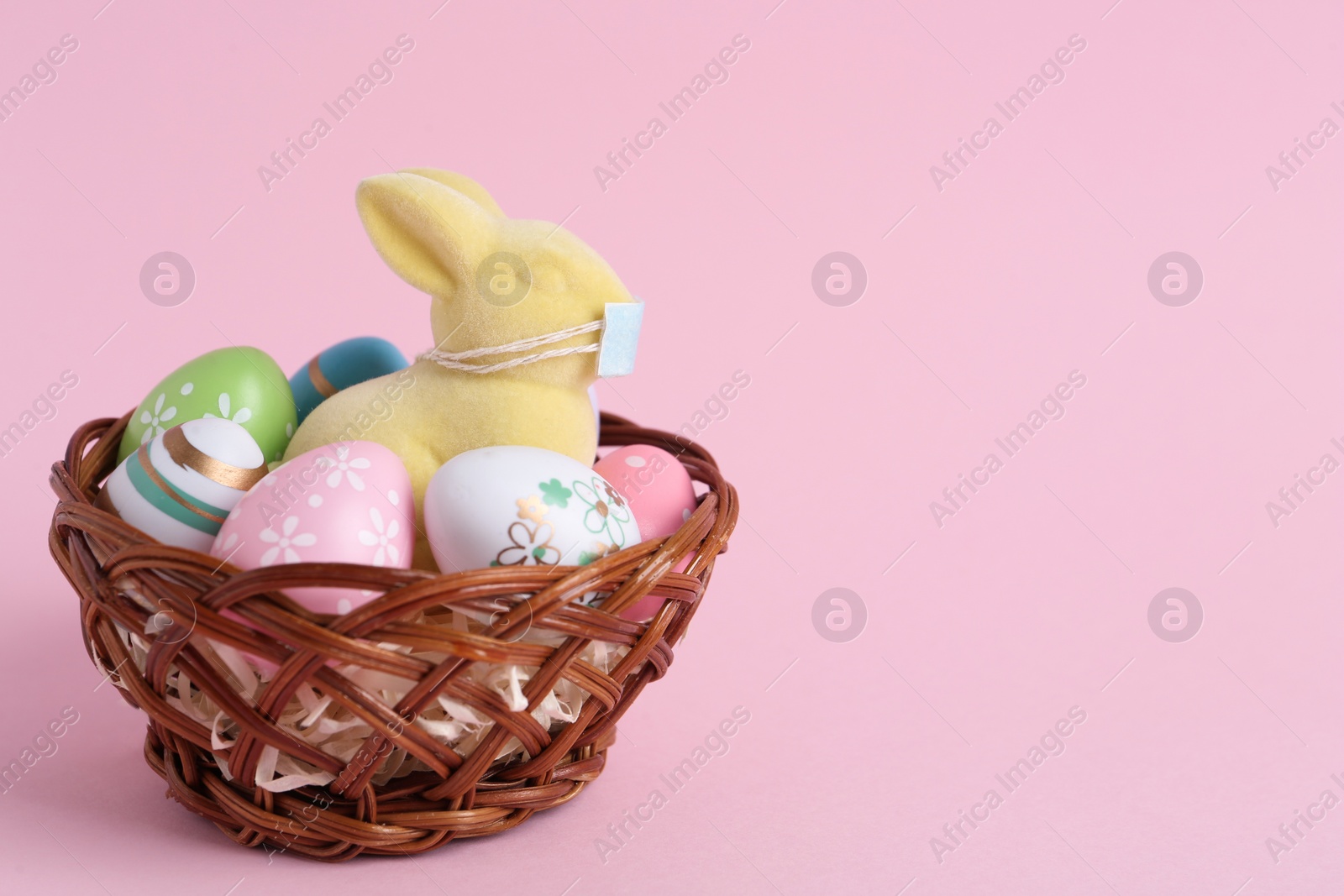 Photo of Cute bunny figure with protective mask and eggs in wicker basket on pink background, space for text. Easter holiday during COVID-19 quarantine