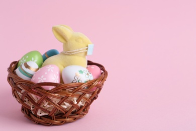 Photo of Cute bunny figure with protective mask and eggs in wicker basket on pink background, space for text. Easter holiday during COVID-19 quarantine