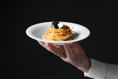 Photo of Waiter holding plate of tasty spaghetti with tomato sauce and black caviar on dark background, closeup. Exquisite presentation of pasta dish