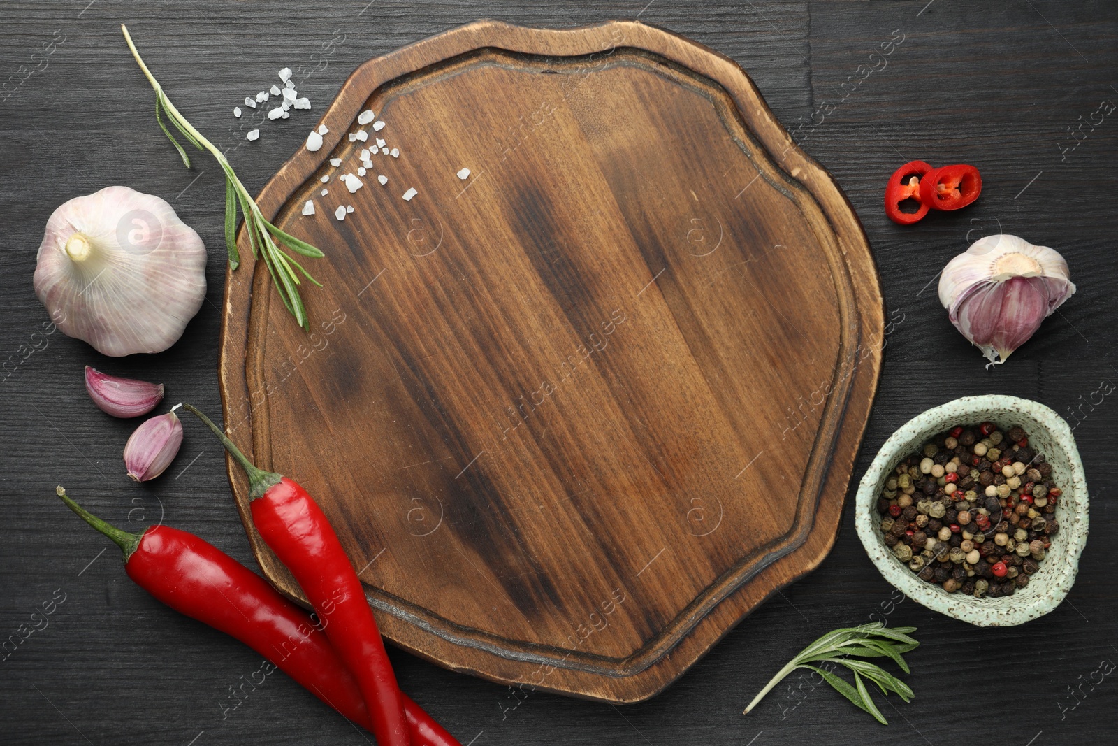 Photo of Cutting board, spices, garlic, rosemary and chili peppers on black wooden table, flat lay. Space for text