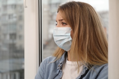 Photo of Sad woman in protective mask near window indoors. Staying at home during coronavirus pandemic