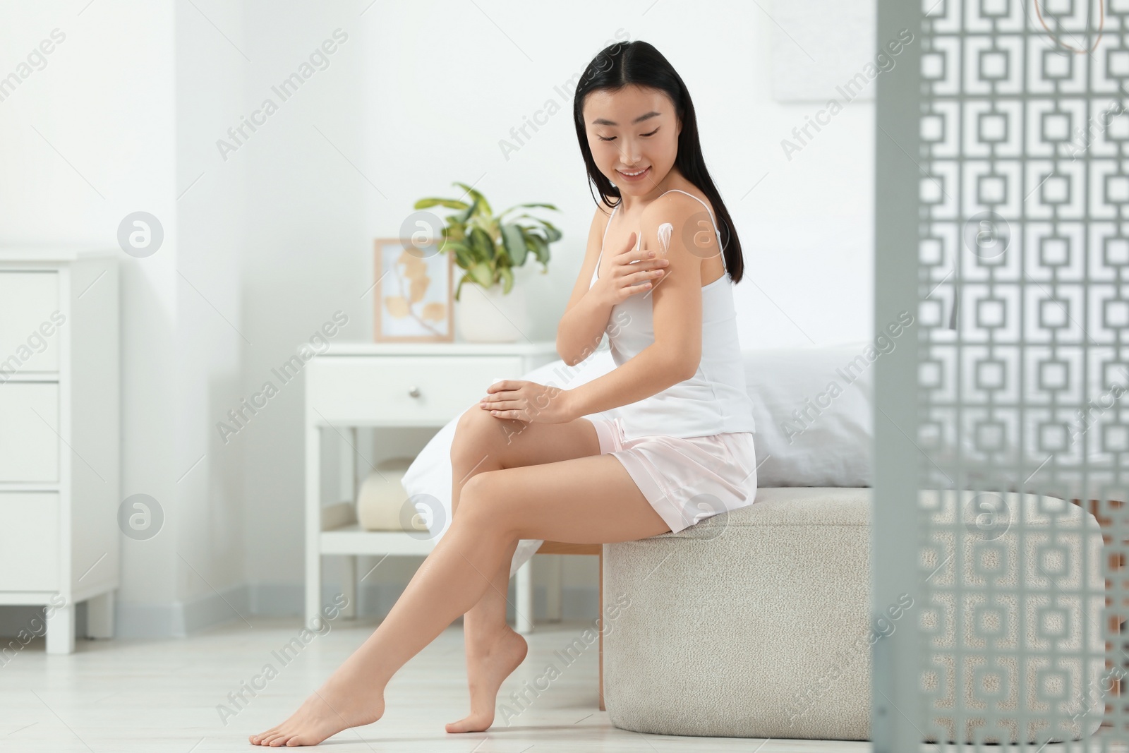 Photo of Beautiful young Asian woman applying body cream on shoulder in bedroom, space for text