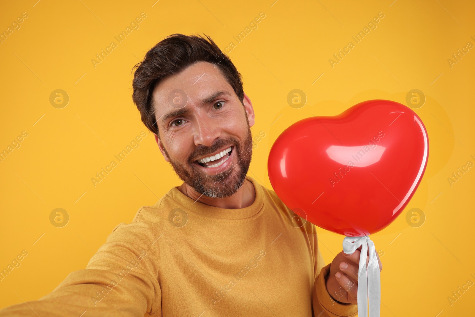 Photo of Happy man holding red heart shaped balloon and taking selfie on orange background