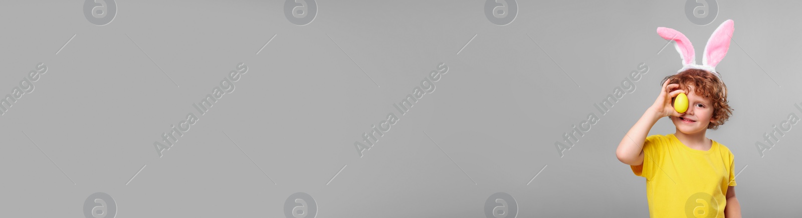 Image of Happy boy with bunny ears holding Easter egg near eye on light grey background. Banner design