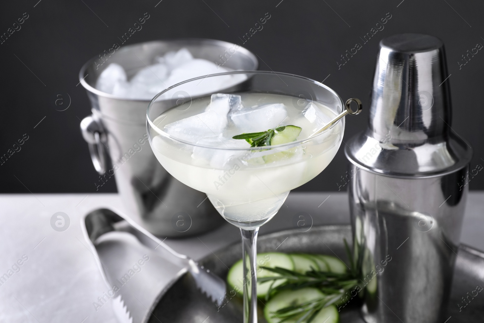 Photo of Glass of martini with cucumber, shaker and ice bucket on table against dark background