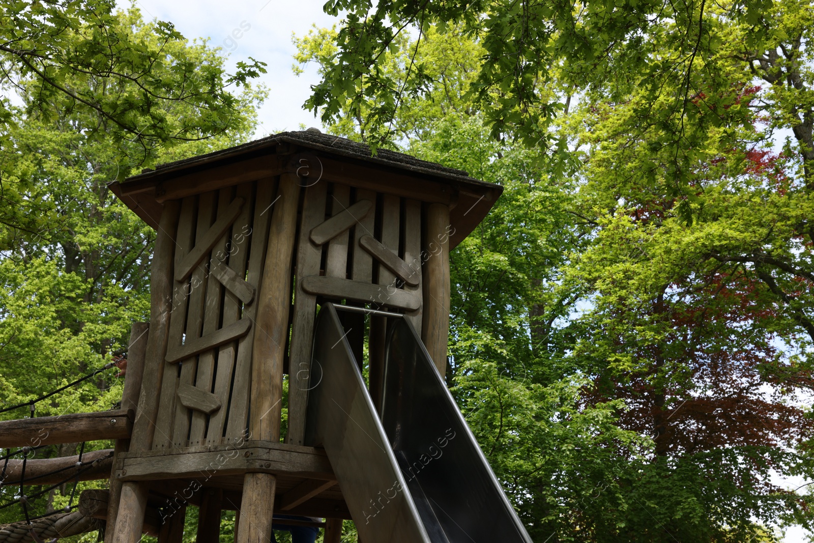 Photo of Wooden slide with bridge near big trees in park. Outdoor playground