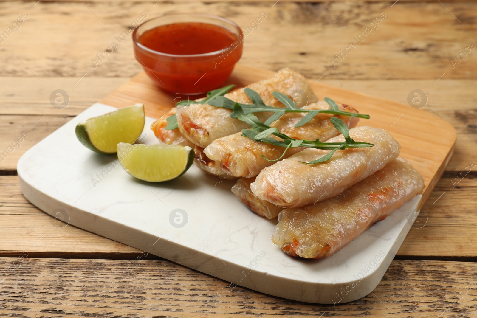 Photo of Tasty fried spring rolls, lime, arugula and sauce on wooden table, closeup