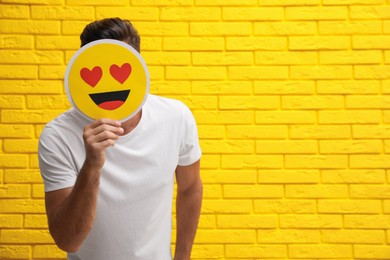Man hiding emotions using card with drawn smiling face near yellow brick wall. Space for text